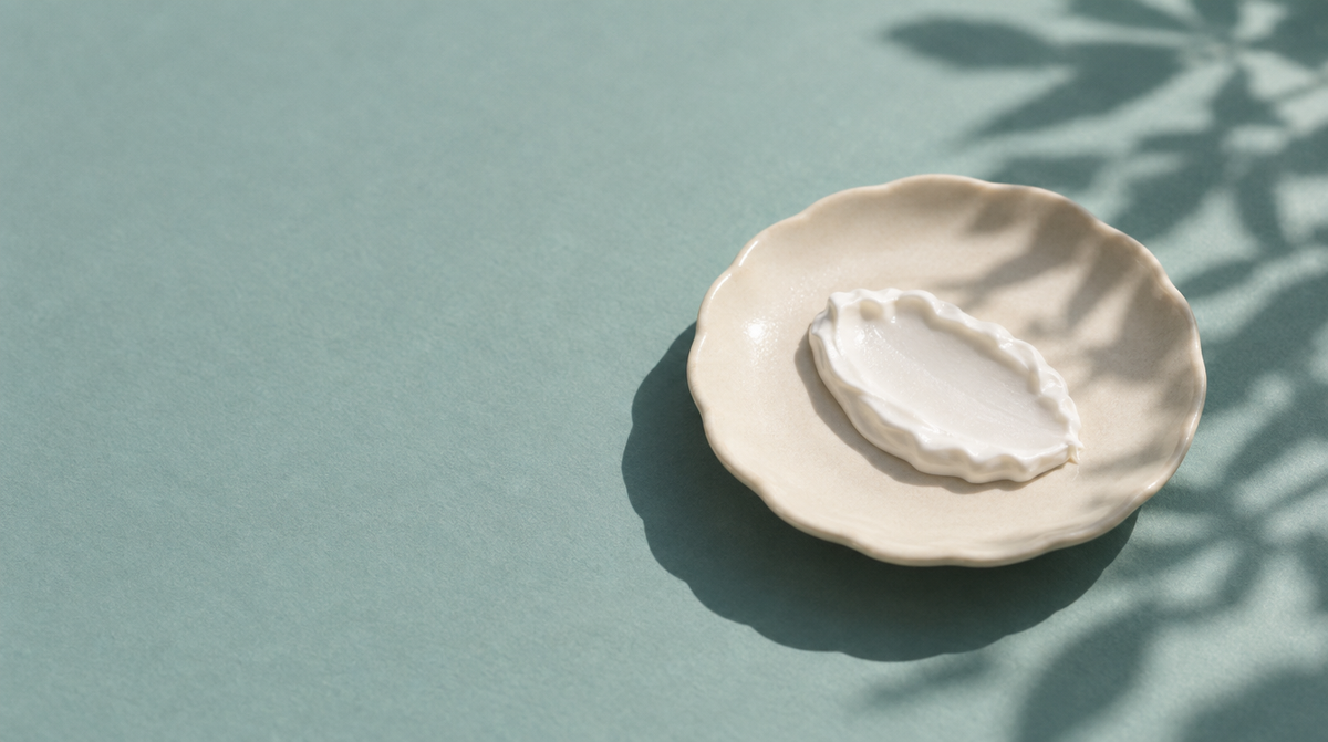 A dollop of thick white mineral sunscreen on a cream ceramic dish, with the shadow of a leaf cast across it in bright daylight.