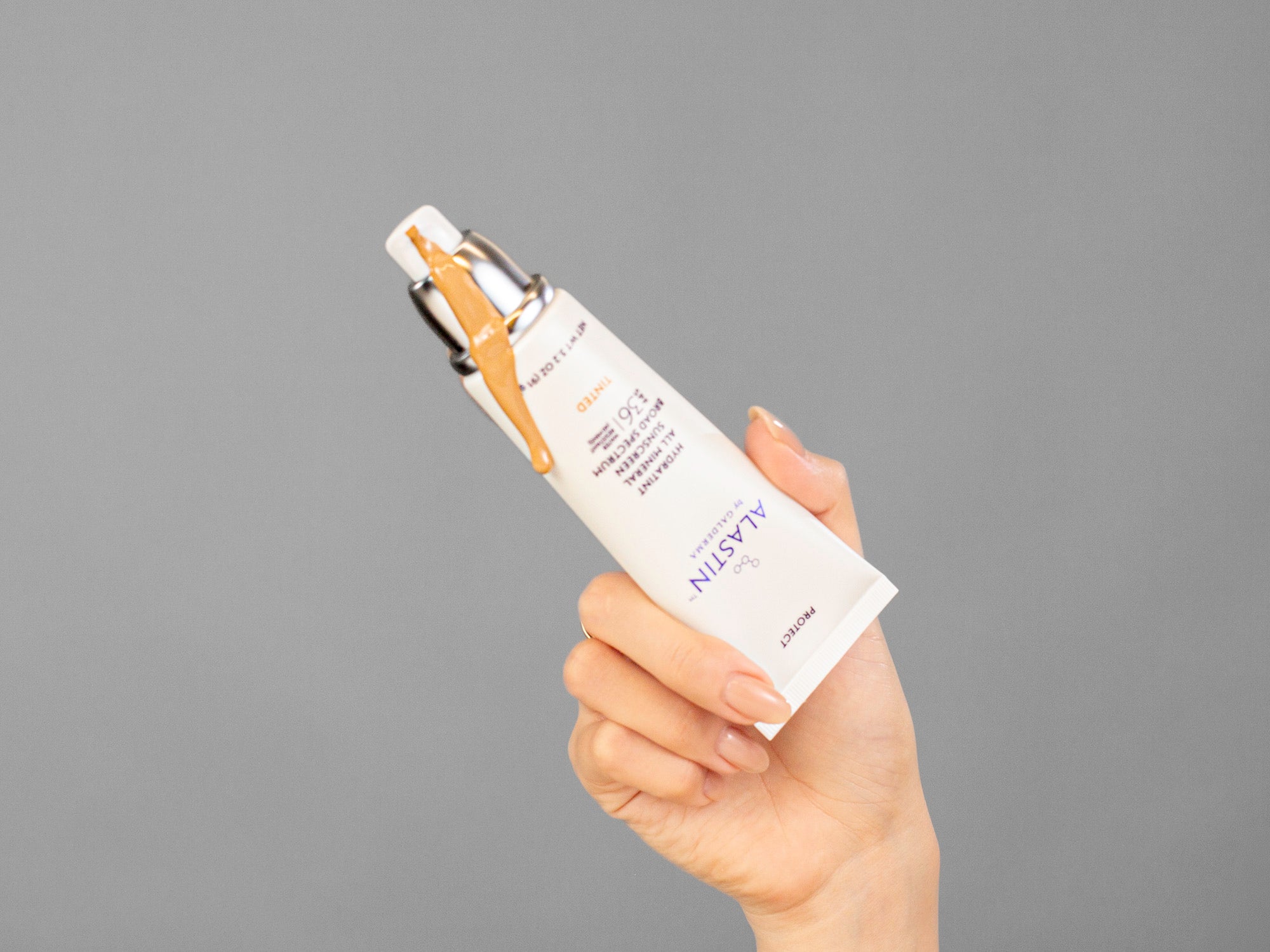 Laziza from LAZ Skincare's hand holding a tube of Alastin Hydratint Pro Mineral Sunscreen SPF36 against a grey background