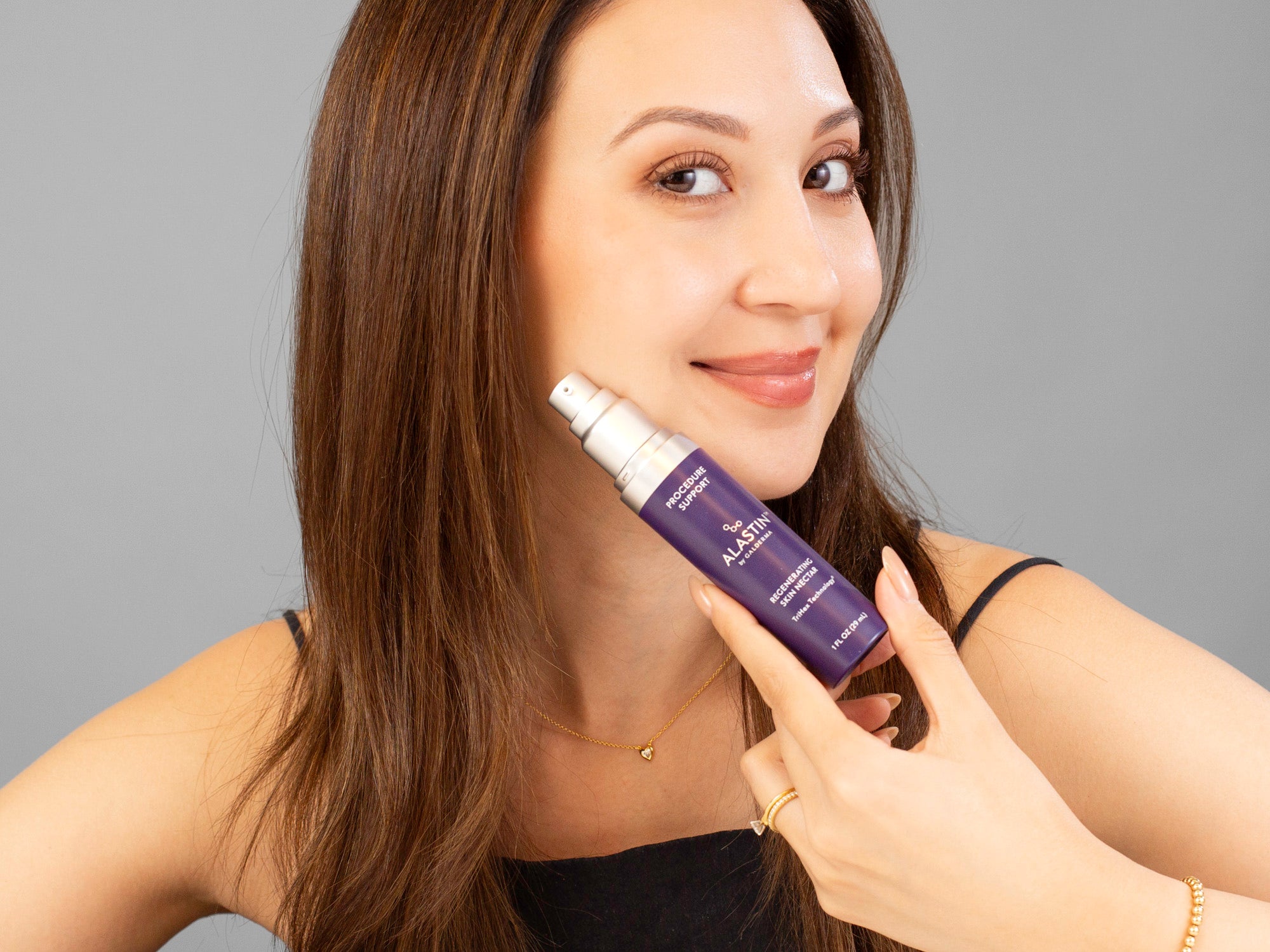 Laziza from LAZ Skincare holding a purple Alastin Regenerating Skin Nectar bottle with a gray background