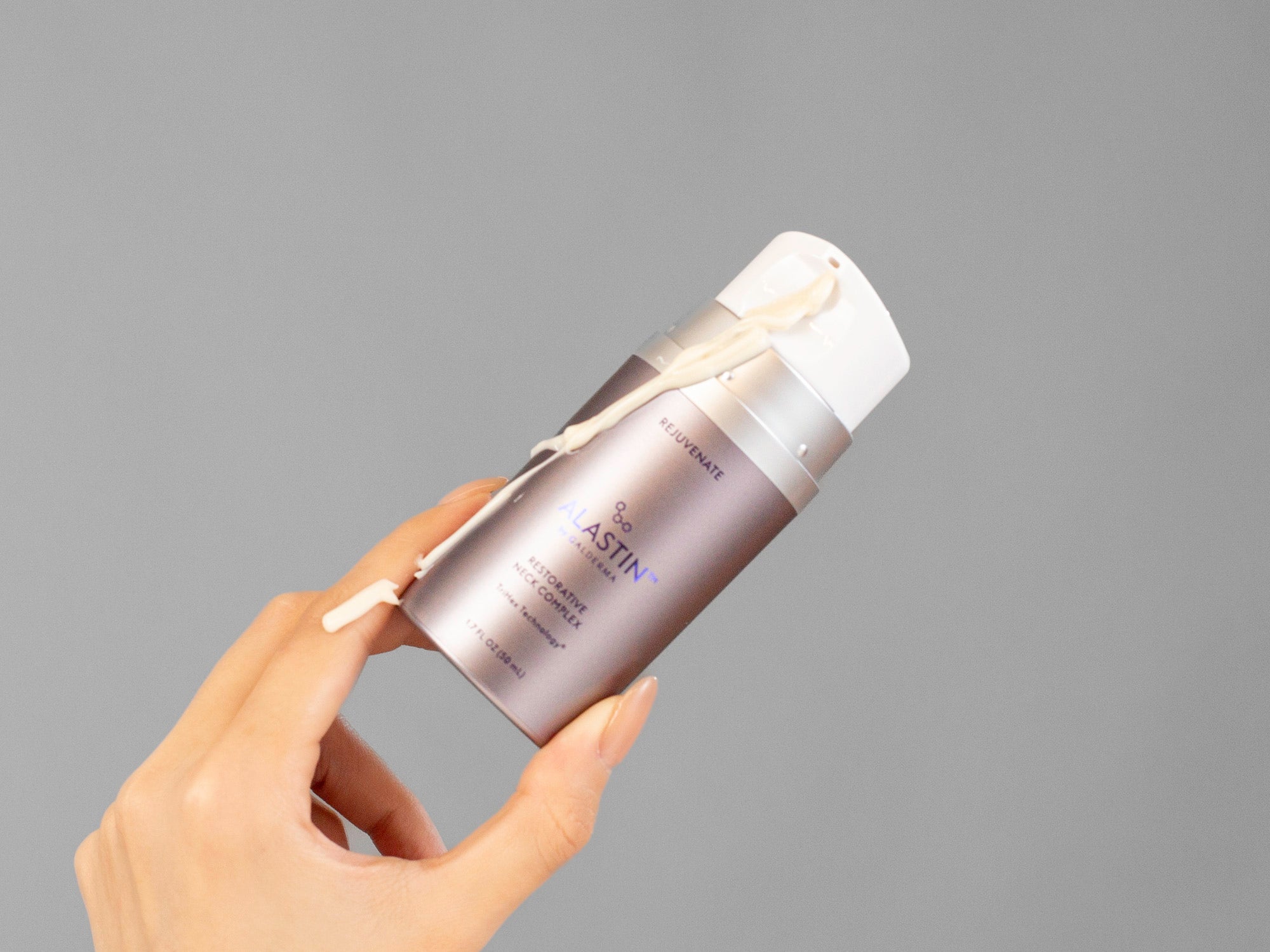 Laziza from LAZ Skincare's hand holding a bottle of Alastin Restorative Neck Complex with a gray background