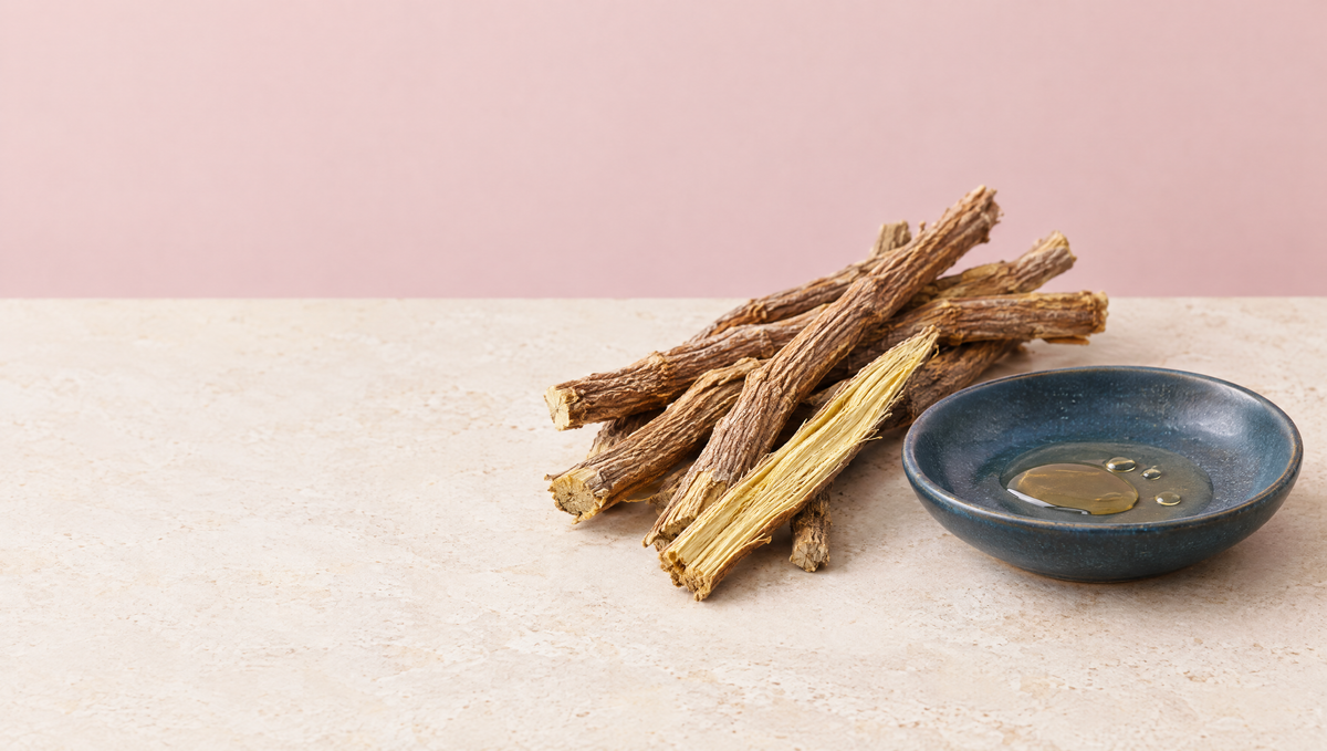 Dried licorice root sticks, one split to show its yellow interior, beside a slate blue dish of pale amber extract on warm stone.