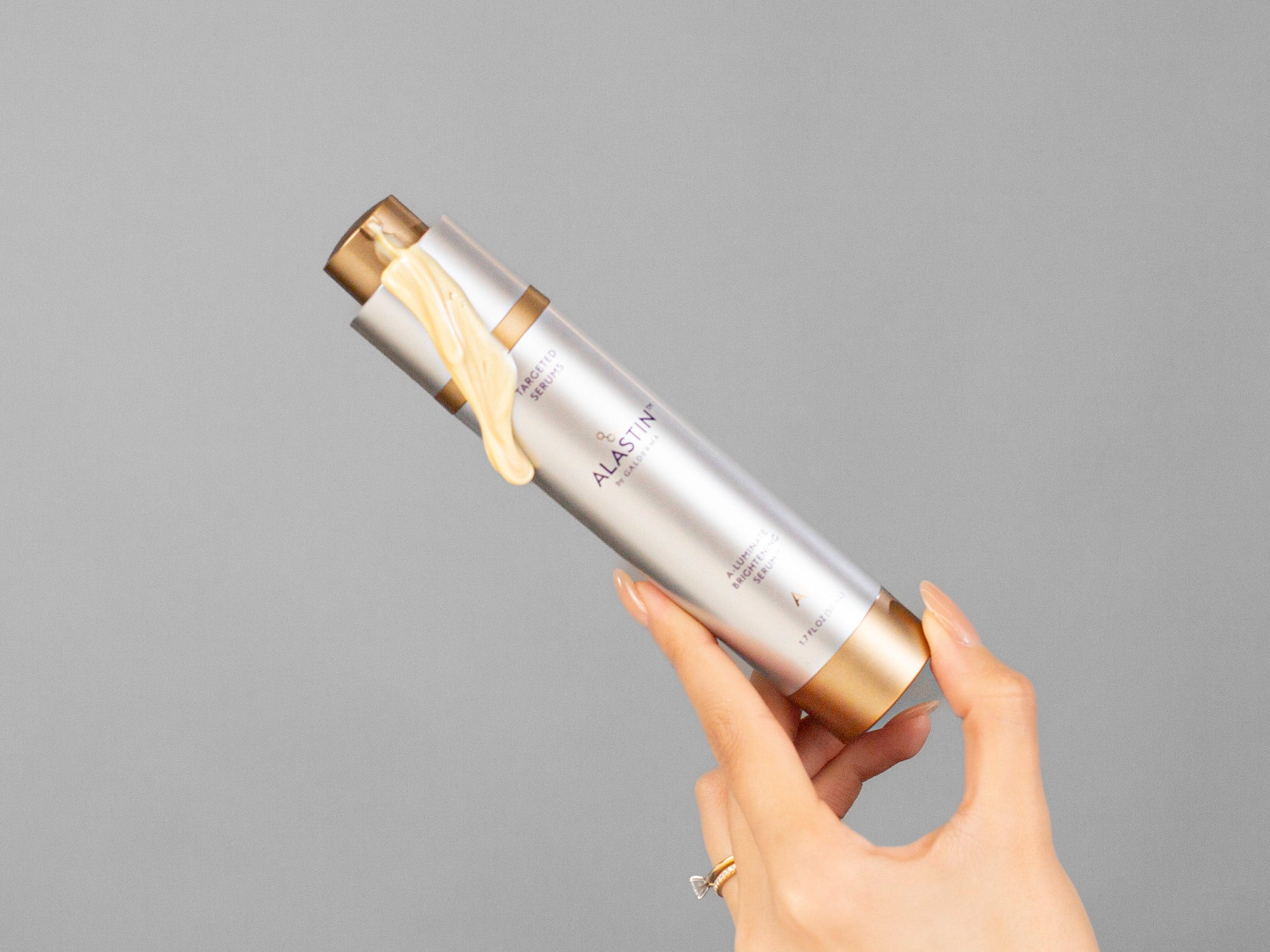 Laziza from LAZ Skincare's hand holding a cylindrical Alastin A-Luminate Brightening Serum bottle with a gray background