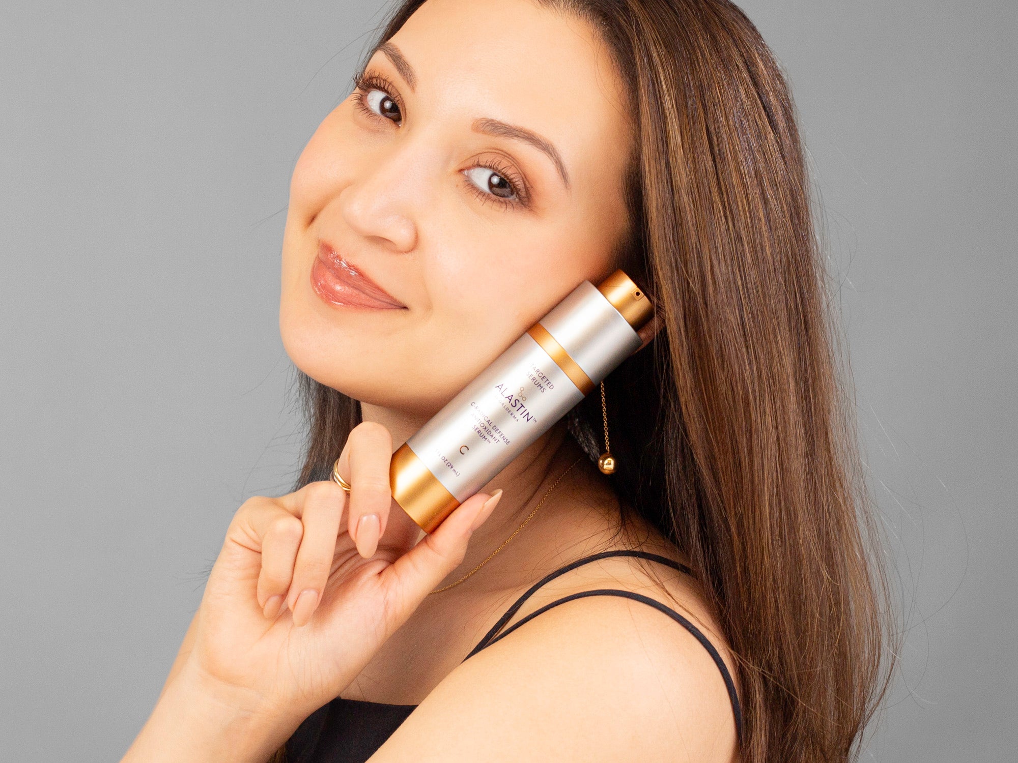 Laziza from LAZ Skincare holding a bottle of lastin C-Radical Defense Antioxidant Serum against a gray background