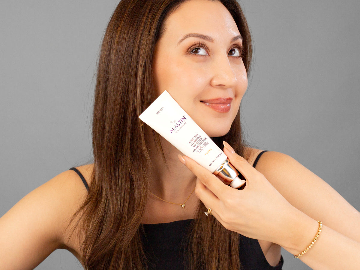 Laziza from LAZ Skincare holding a bottle of Alastin Hydratint Pro Mineral Sunscreen SPF 36 up to her face
