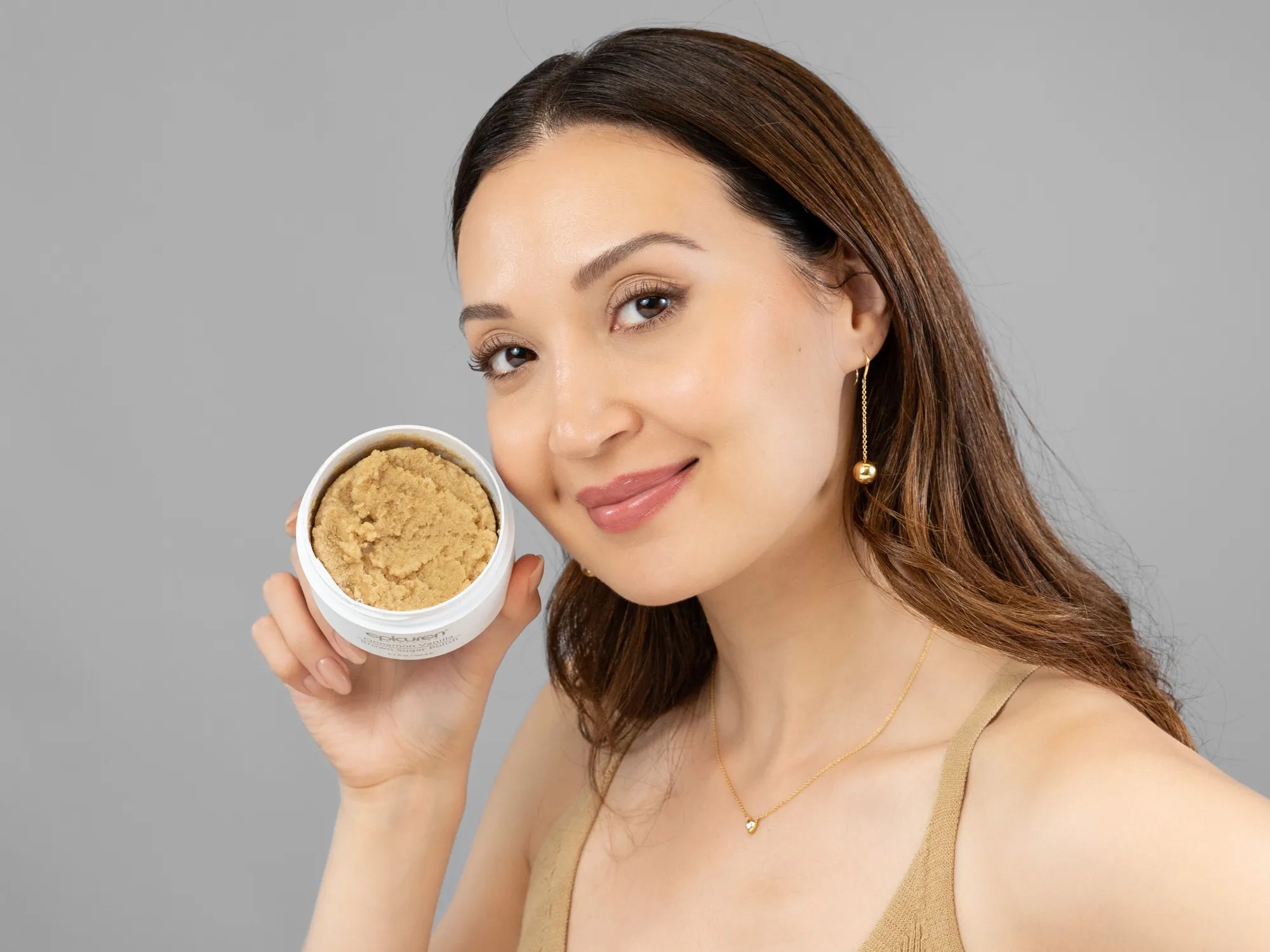 Laziza from LAZ Skincare holding a bottle of Epicuren Cinnamon Vanilla Brown Sugar Polish up to her face