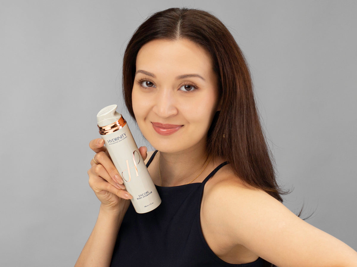 Laziza from LAZ Skincare holding a bottle of Hydrinity Encore Body Hydrator up to her face